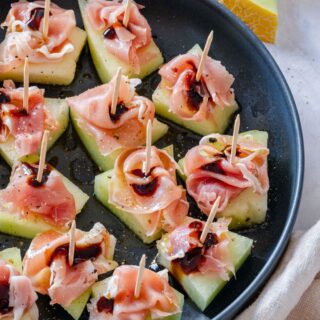 Prosciutto and melon appetizers with balsamic glaze on a black plate.