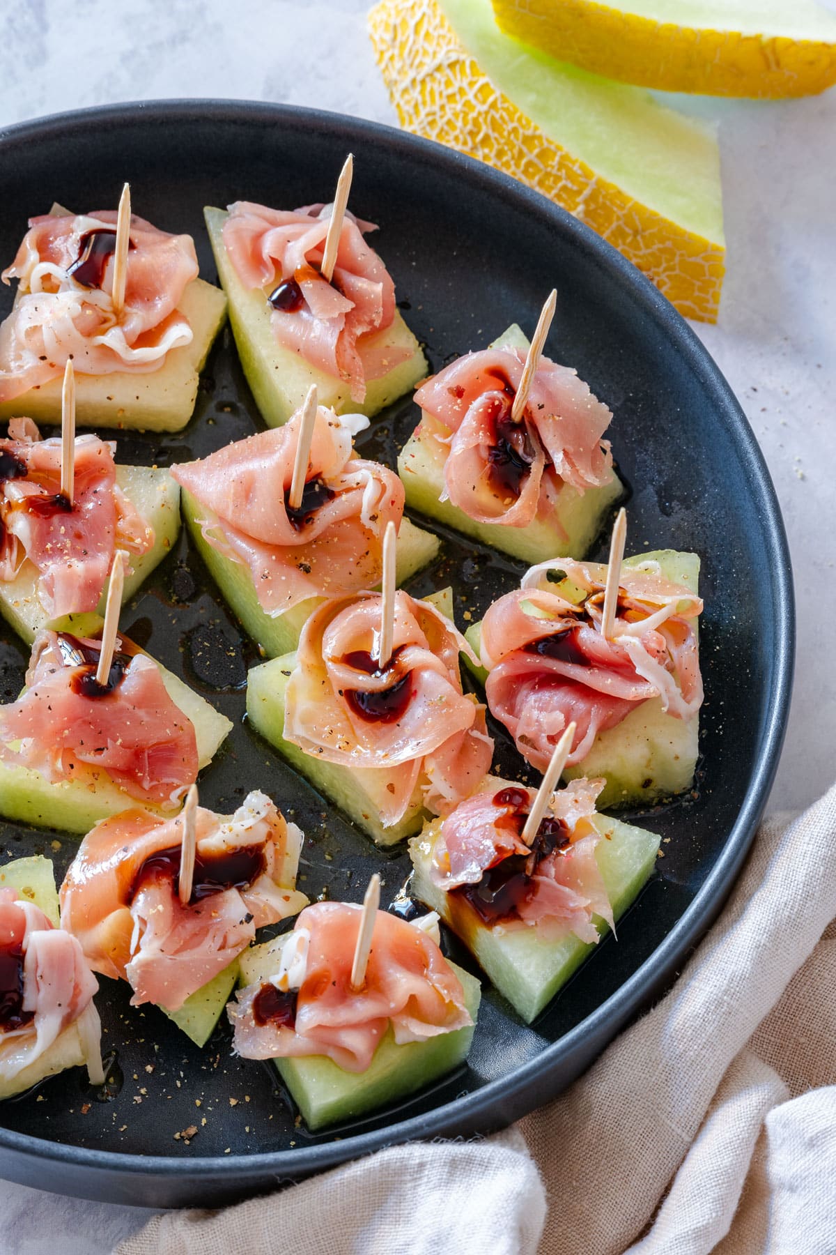 Prosciutto and melon appetizers with balsamic glaze on a black plate.