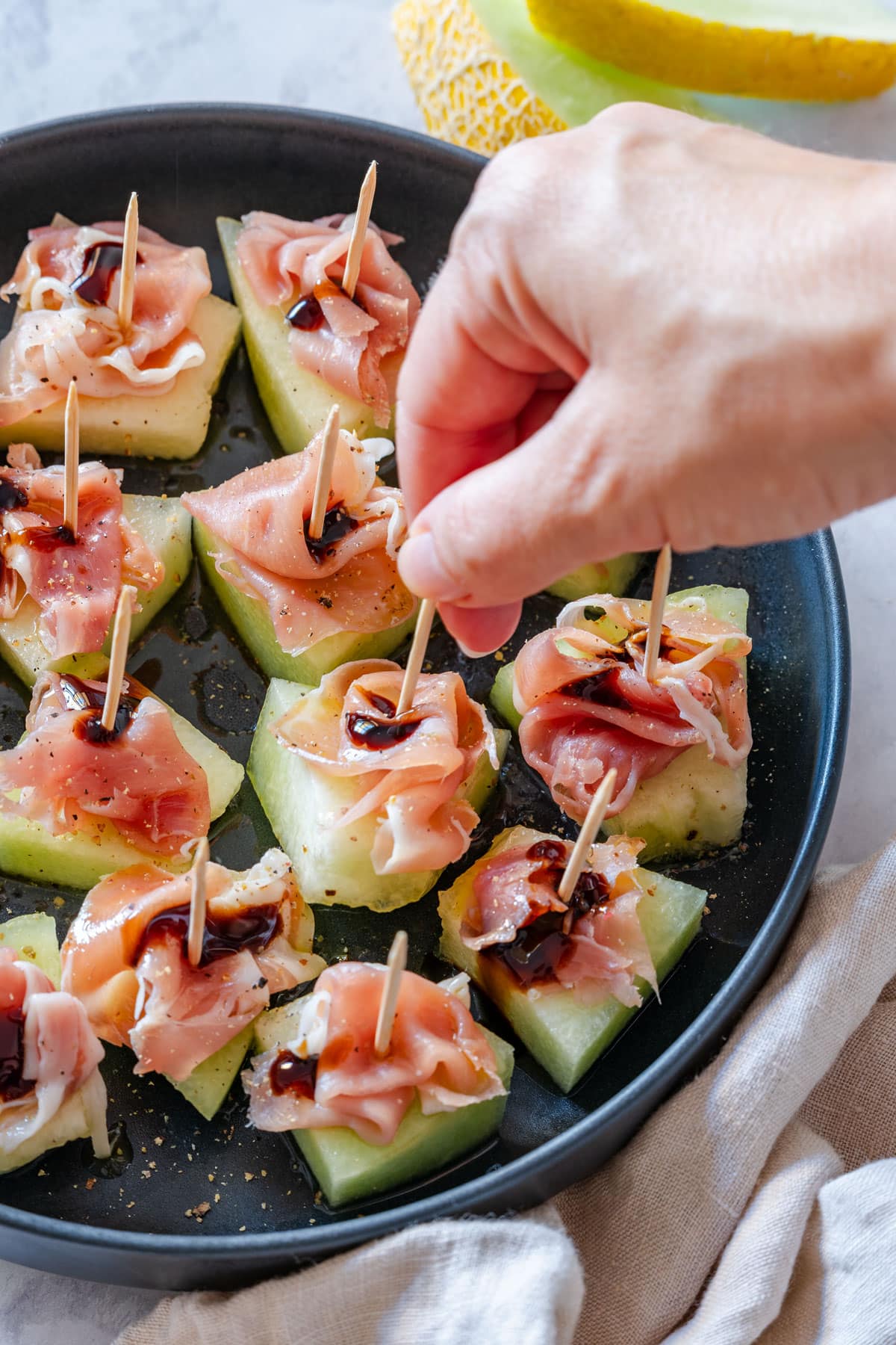 A hand reaching for a melon with prosciutto, topped with a drizzle of balsamic glaze.