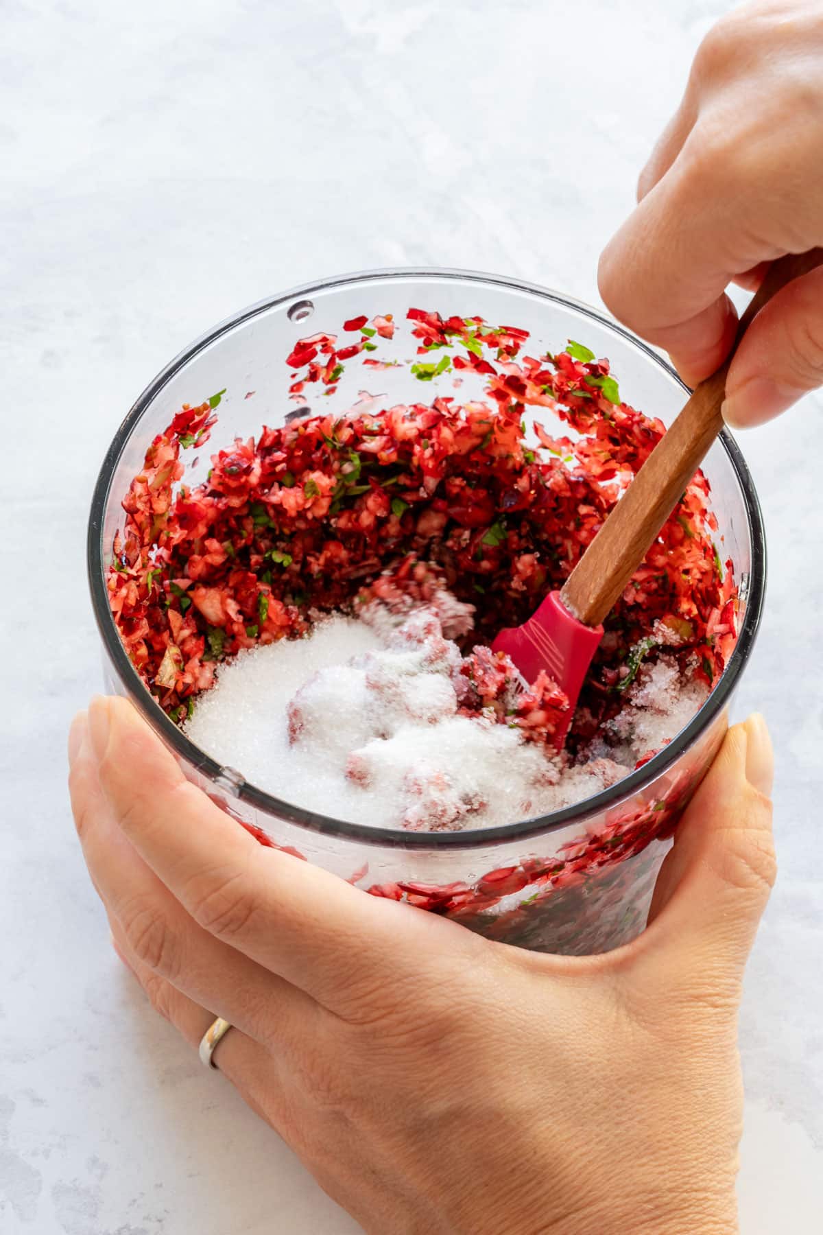 Julia is mixing chopped cranberries and herbs with sugar in a glass bowl using a spatula.