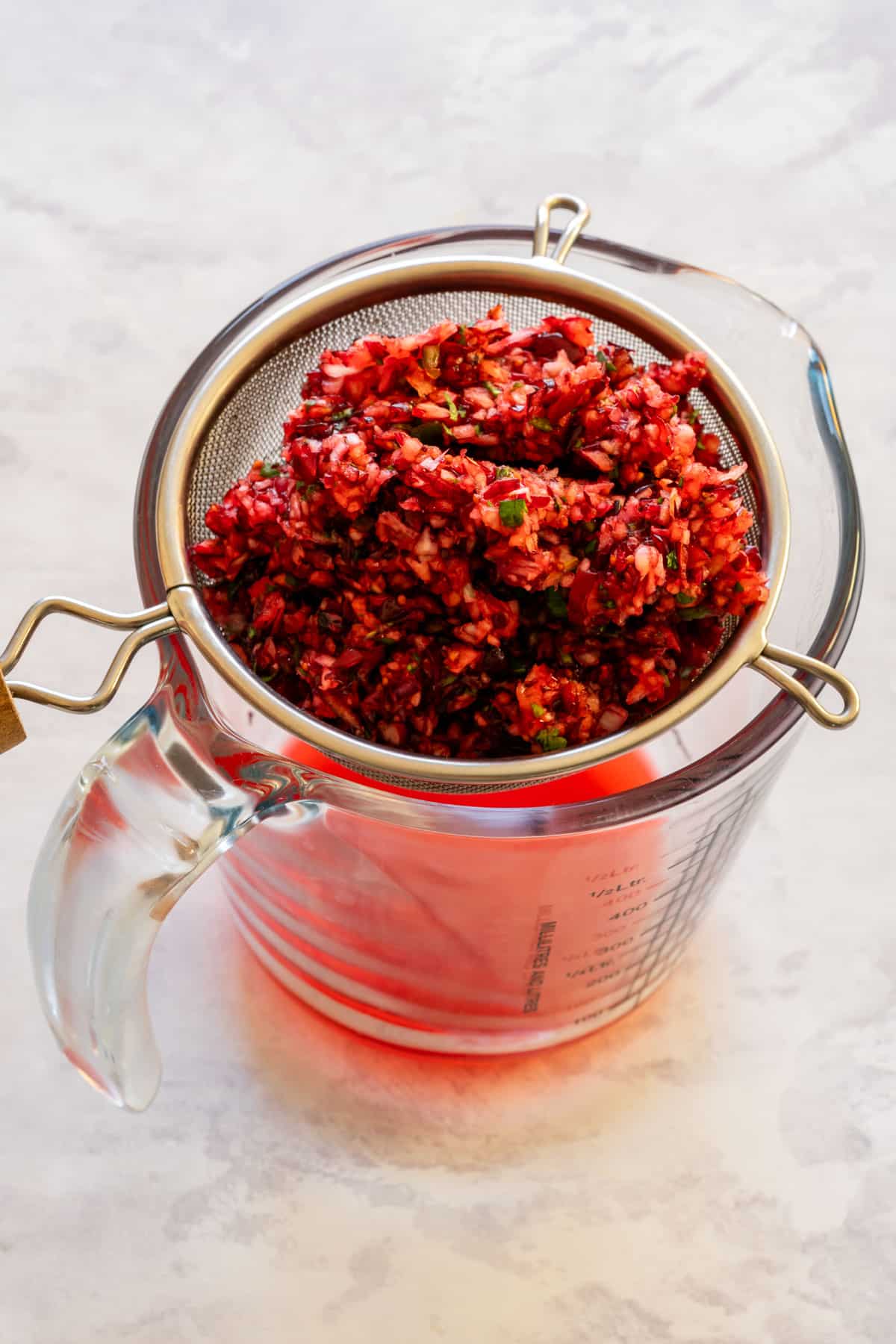 A strainer with cranberry mixture over a glass measuring cup collecting red liquid.