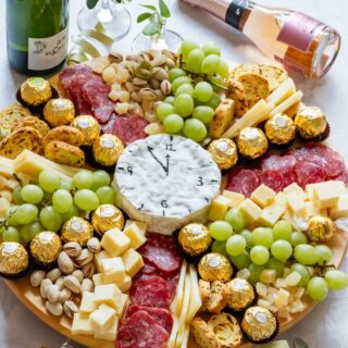 Festive charcuterie board with grapes, cheese cubes, salami, pistachios, chocolates, and a clock-shaped cheese.