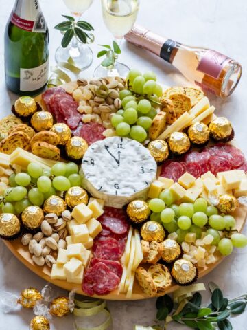 Festive charcuterie board with grapes, cheese cubes, salami, pistachios, chocolates, and a clock-shaped cheese.