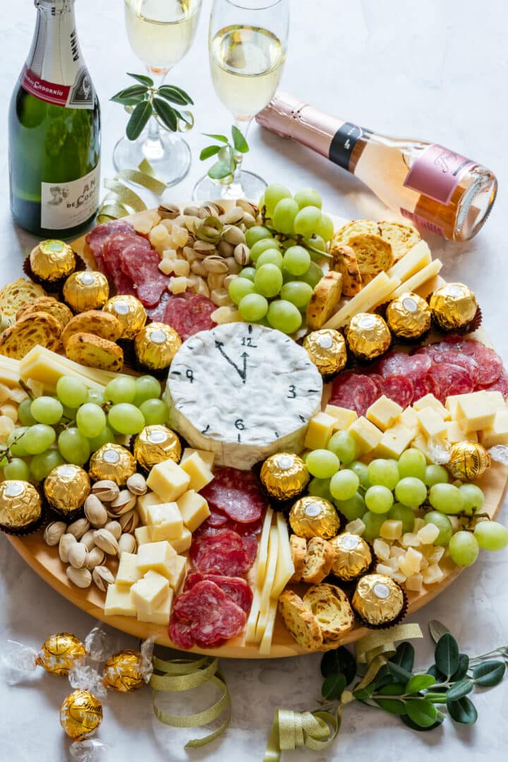 Festive charcuterie board with grapes, cheese cubes, salami, pistachios, chocolates, and a clock-shaped cheese.