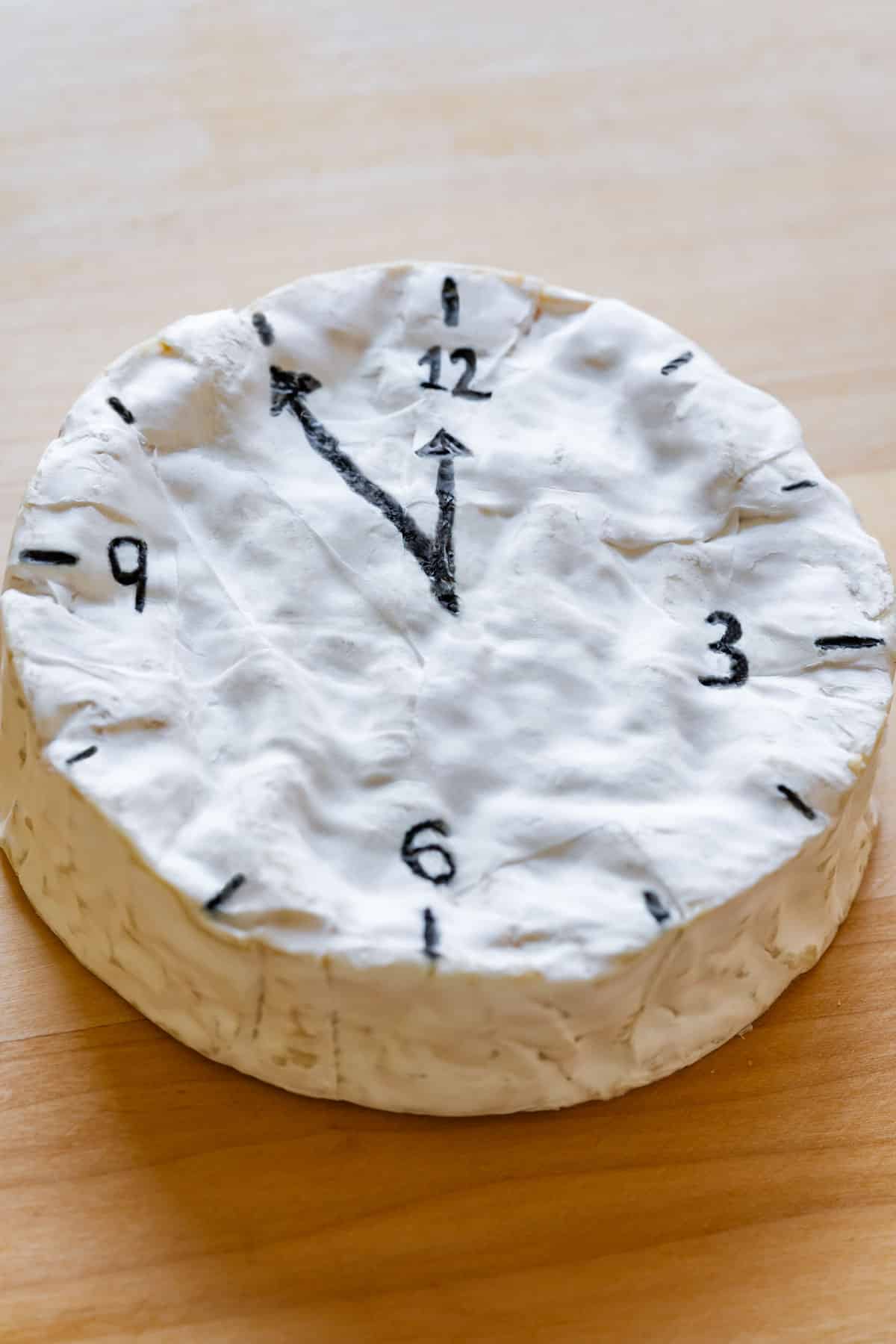 Camembert cheese with black clock markings and hands drawn on its surface.