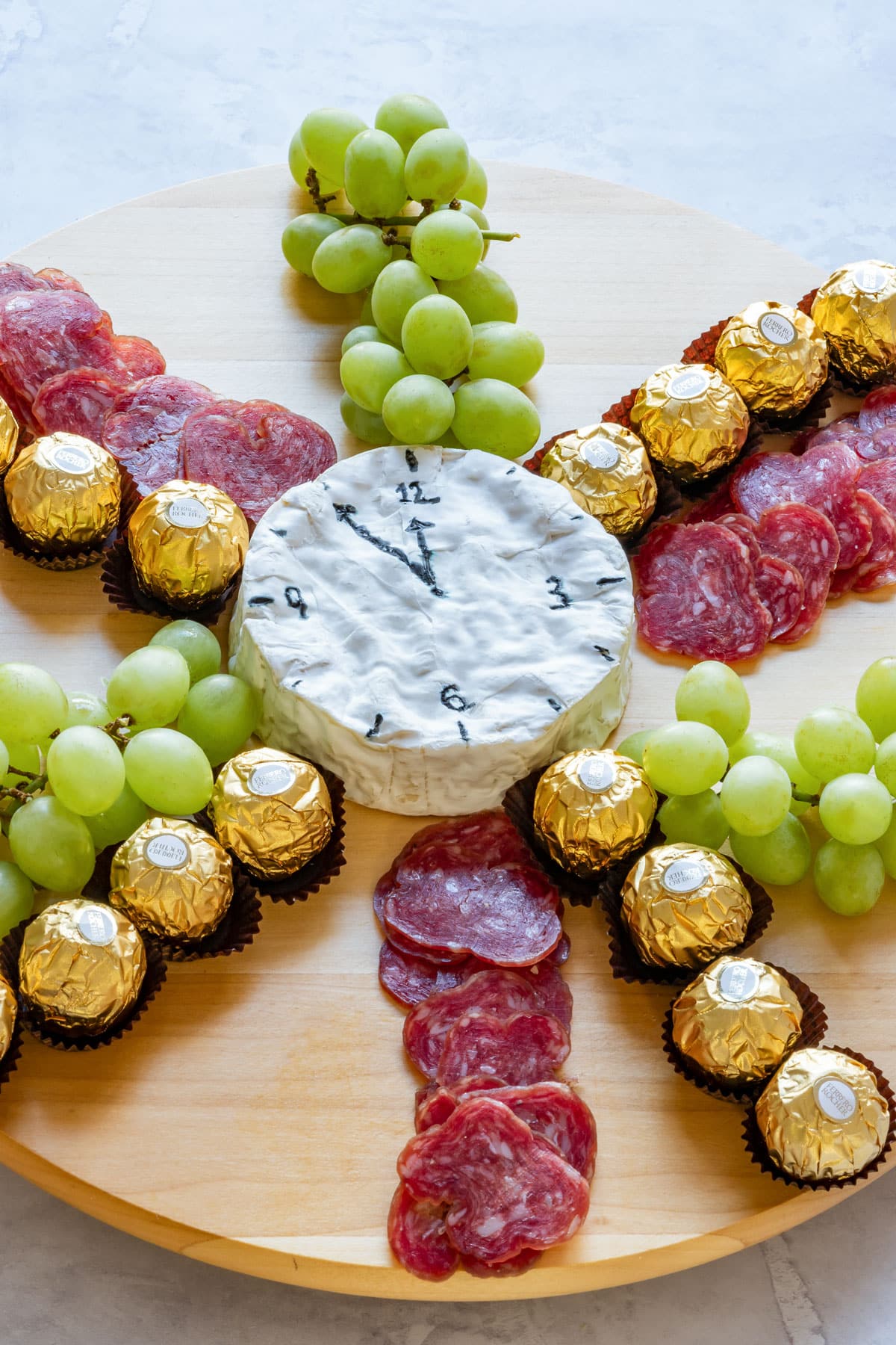 Round Camembert cheese wheel designed as a clock, surrounded by grapes, salami and chocolates on a wooden board.