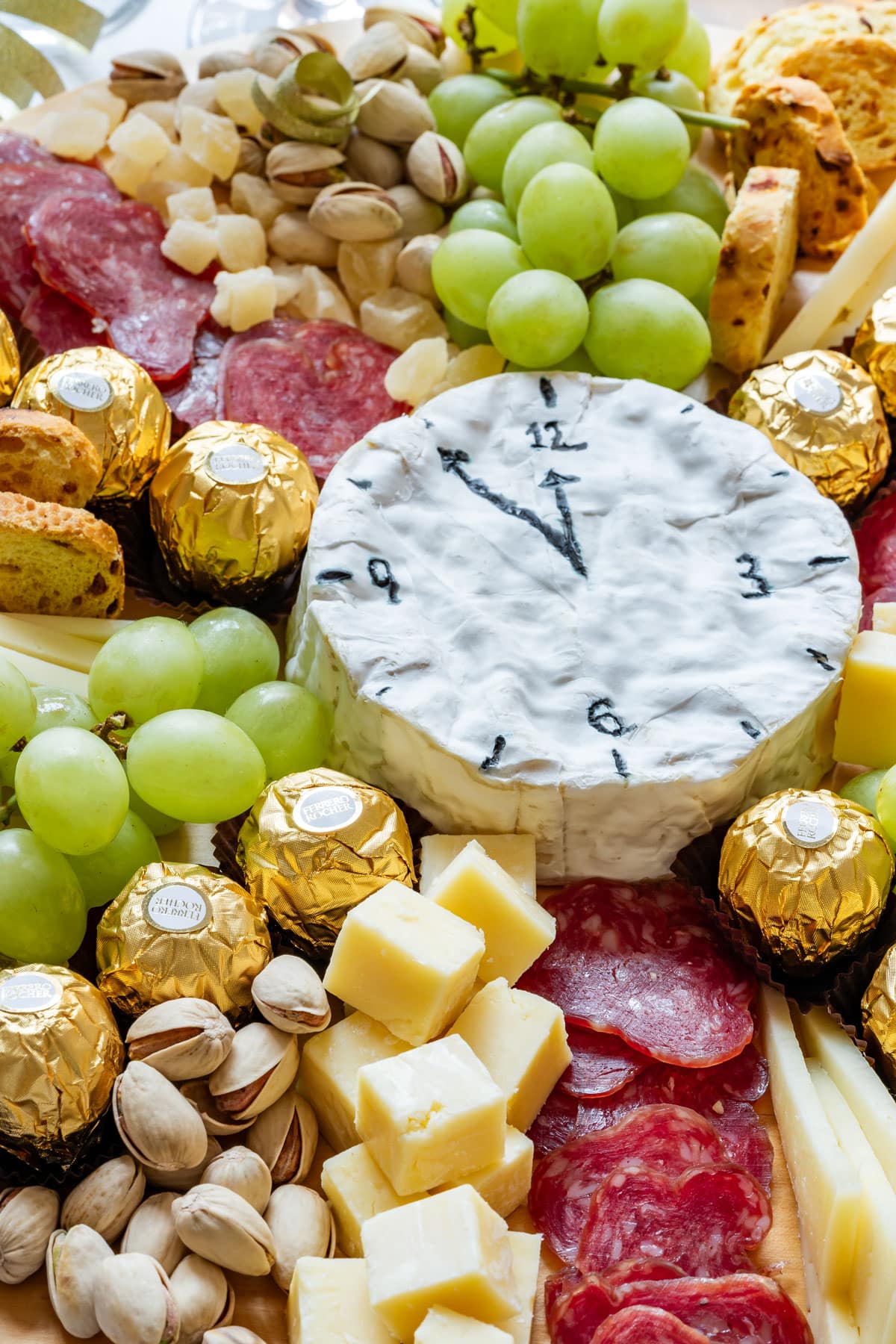 Cheese clock surrounded by grapes, chocolates, nuts, salami and cheese cubes on a charcuterie board.