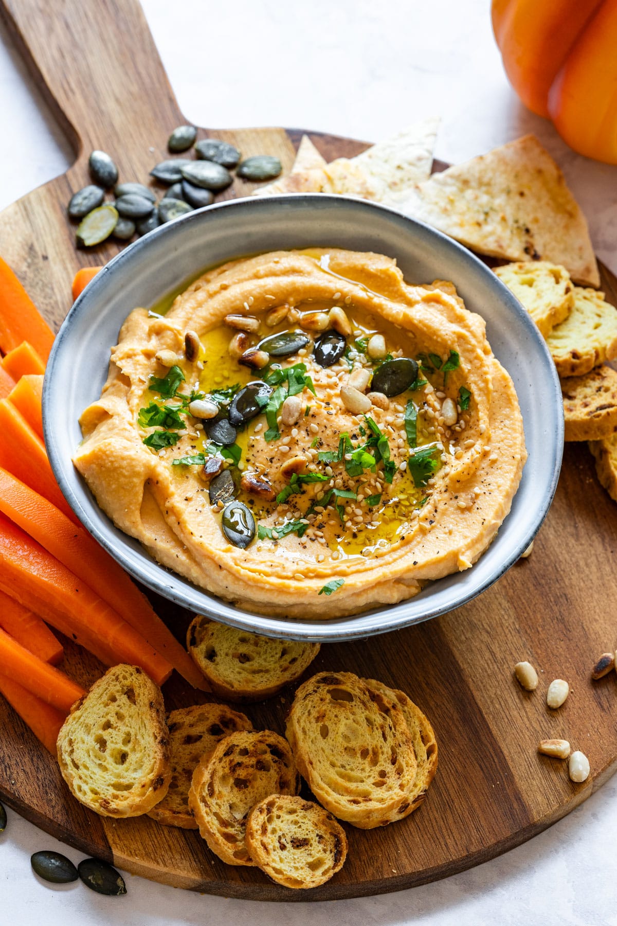 Bowl of hummus is surrounded by carrot sticks, toasted bread slices and tortilla chips on a wooden serving board.