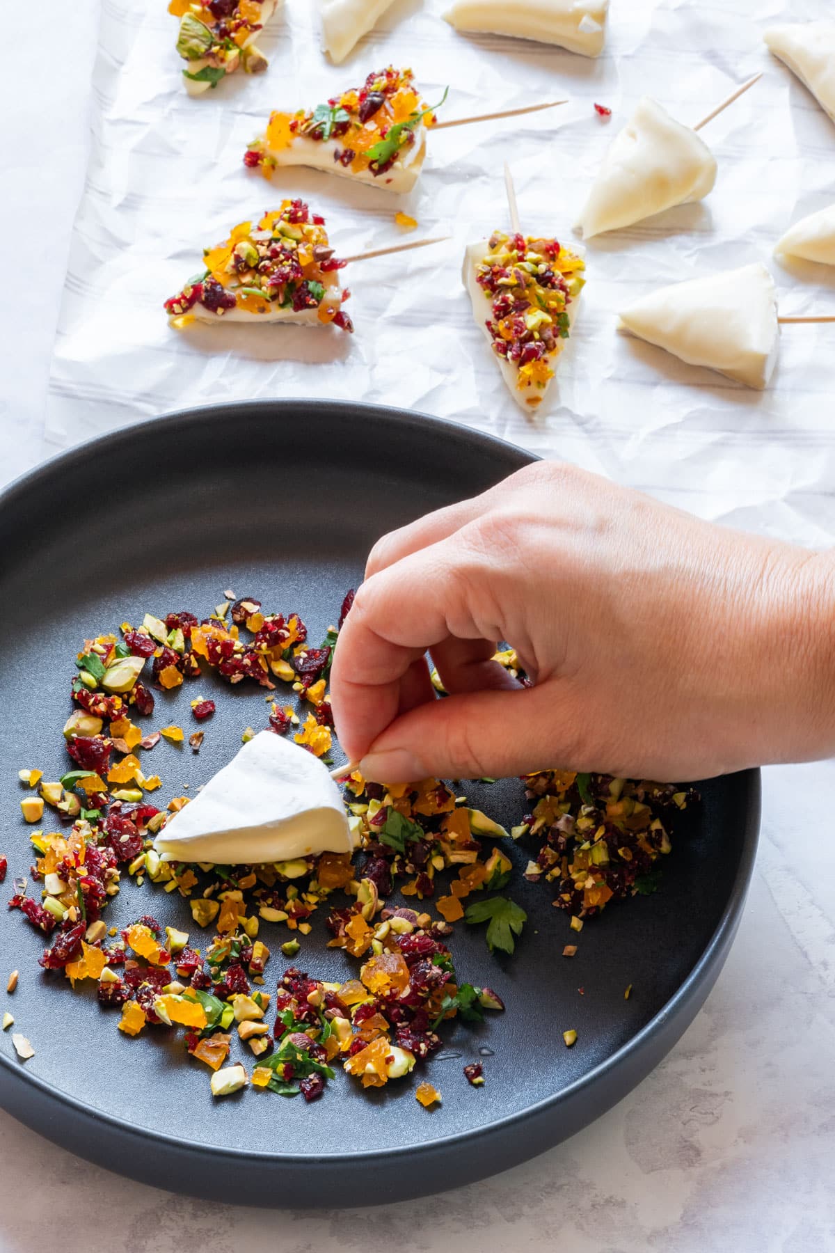 A hand dips a brie cheese wedge into a mix of chopped nuts and dried fruits on a black plate.