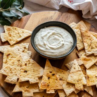 A small bowl of creamy dip surrounded by Christmas tree-shaped tortilla chips on a wooden board.