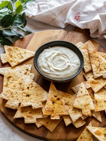 A small bowl of creamy dip surrounded by Christmas tree-shaped tortilla chips on a wooden board.