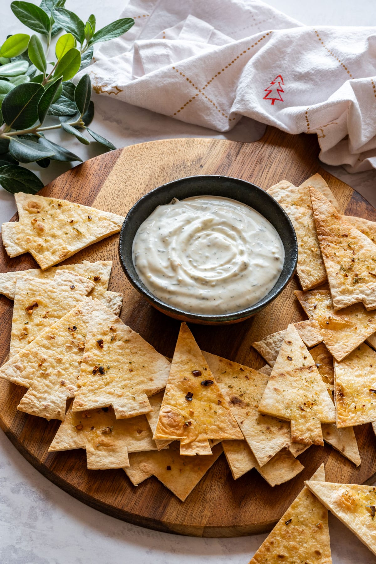 A small bowl of creamy dip surrounded by Christmas tree-shaped tortilla chips on a wooden board.