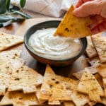 A hand dips a Christmas tree tortilla chip into a bowl of creamy dip, surrounded by more chips on a wooden board.