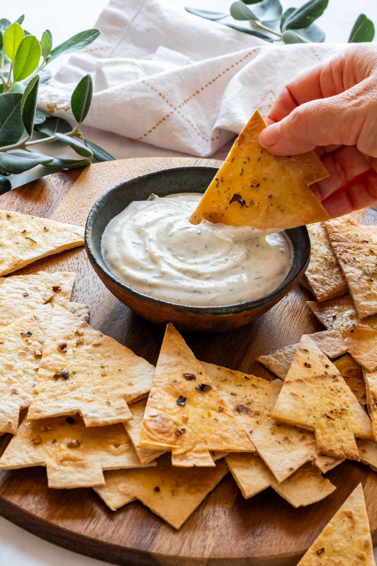 A hand dips a Christmas tree tortilla chip into a bowl of creamy dip, surrounded by more chips on a wooden board.