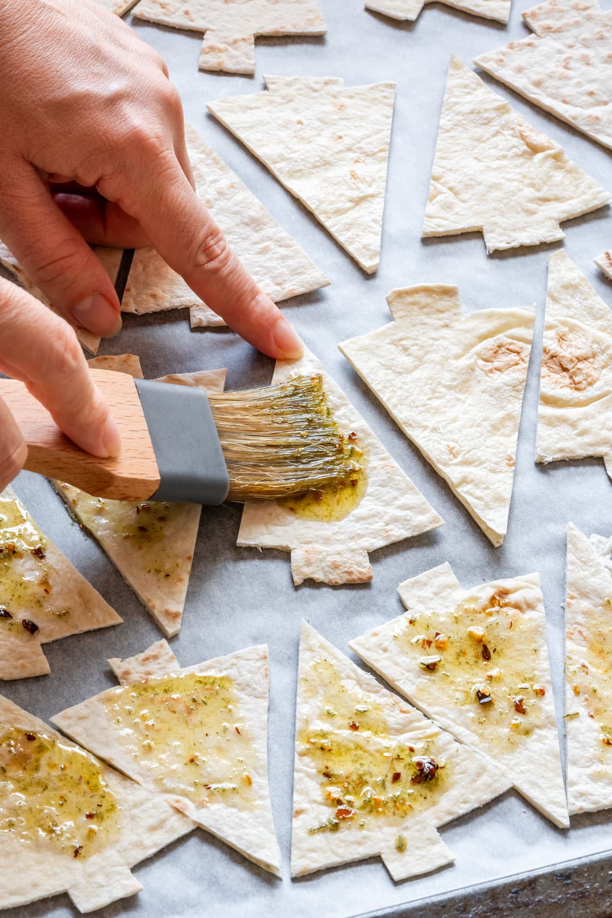 Brushing seasoned oil onto Christmas tree-shaped tortilla pieces on a baking sheet.