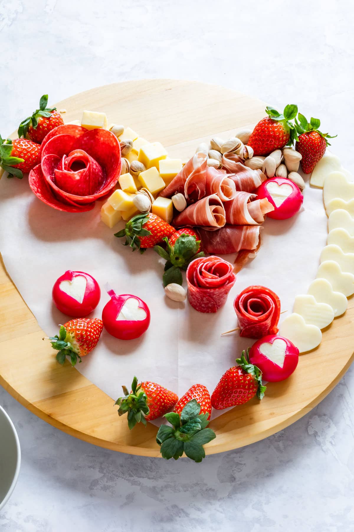 Assembling the heart-shaped charcuterie board with salami roses, a variety of meats and cheeses strawberries, and nuts.