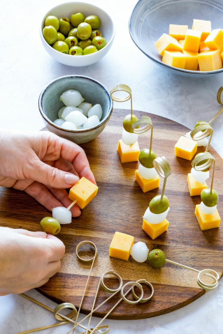 Julia assembling appetizer skewers with cheddar cheese, pearl onions and green olives on a wooden board, with extra ingredients and skewers nearby.