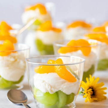 Mini Irish flag dessert cups with green grapes, creamy white layer and mandarin orange segments in clear cups, garnished with a yellow flower and spoon nearby.