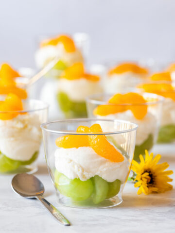 Mini Irish flag dessert cups with green grapes, creamy white layer and mandarin orange segments in clear cups, garnished with a yellow flower and spoon nearby.