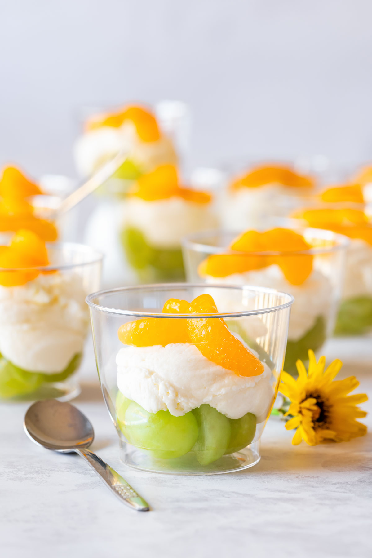 Mini Irish flag dessert cups with green grapes, creamy white layer and mandarin orange segments in clear cups, garnished with a yellow flower and spoon nearby.