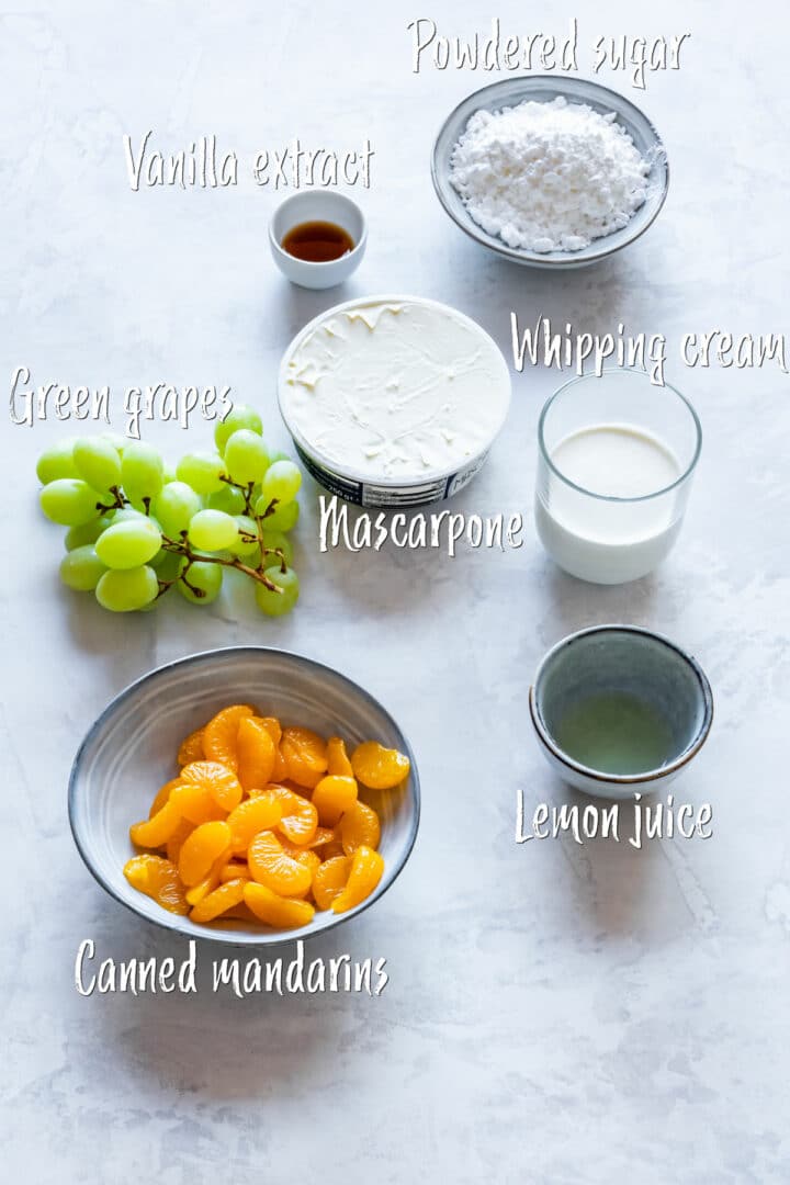 Ingredients for Irish-themed dessert cups displayed on a marble surface: green grapes, canned mandarin oranges, mascarpone, whipping cream, powdered sugar, vanilla extract and lemon juice.