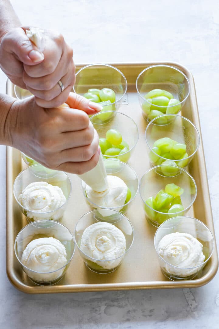 Piping creamy mascarpone mixture over green grapes in clear dessert cups on a baking tray, step in making layered party desserts.