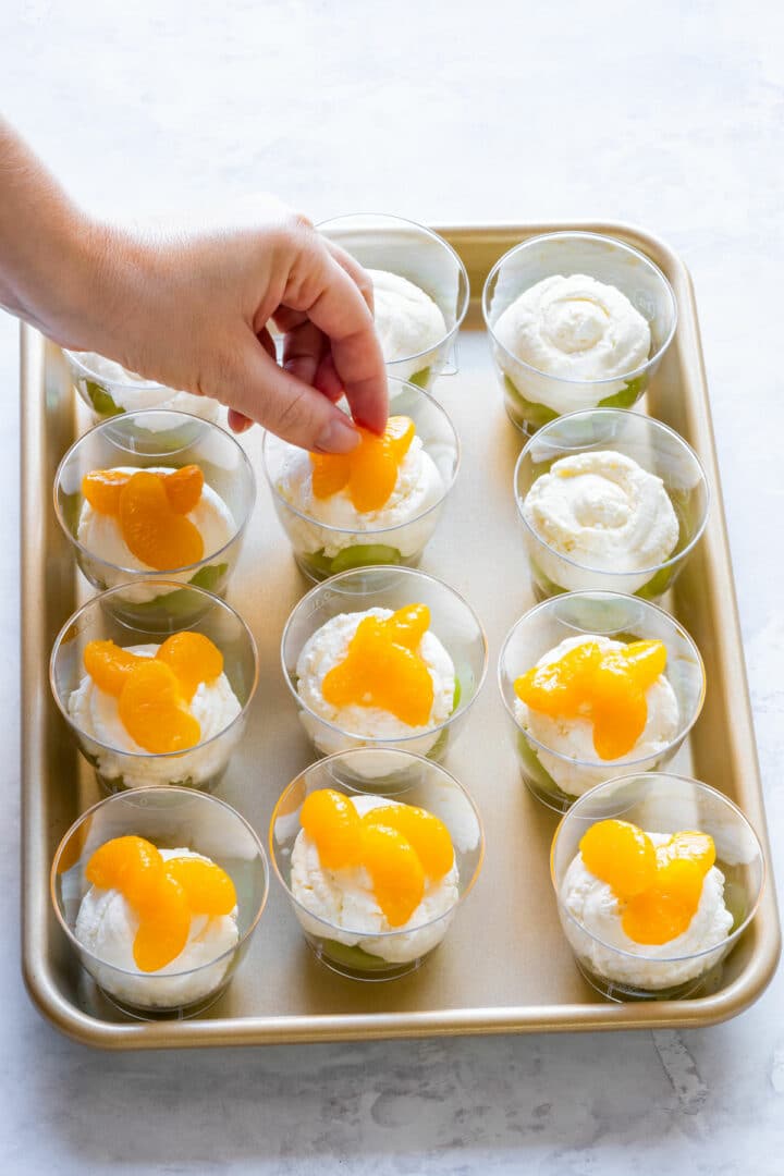 Adding mandarin orange segments to dessert cups with green grapes and a creamy layer, final step in assembling layered party treats.