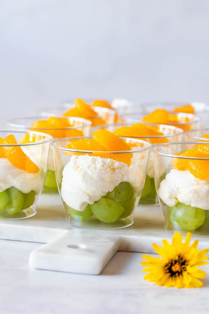 Layered dessert cups with green grapes, creamy white filling and mandarin orange segments, served on a marble board with a yellow flower.