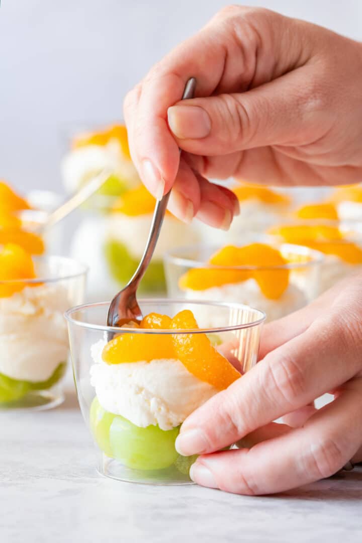 Close-up of spooning a layered party dessert cup with green grapes, creamy mascarpone filling and mandarin orange segments, inspired by Irish flag colors.